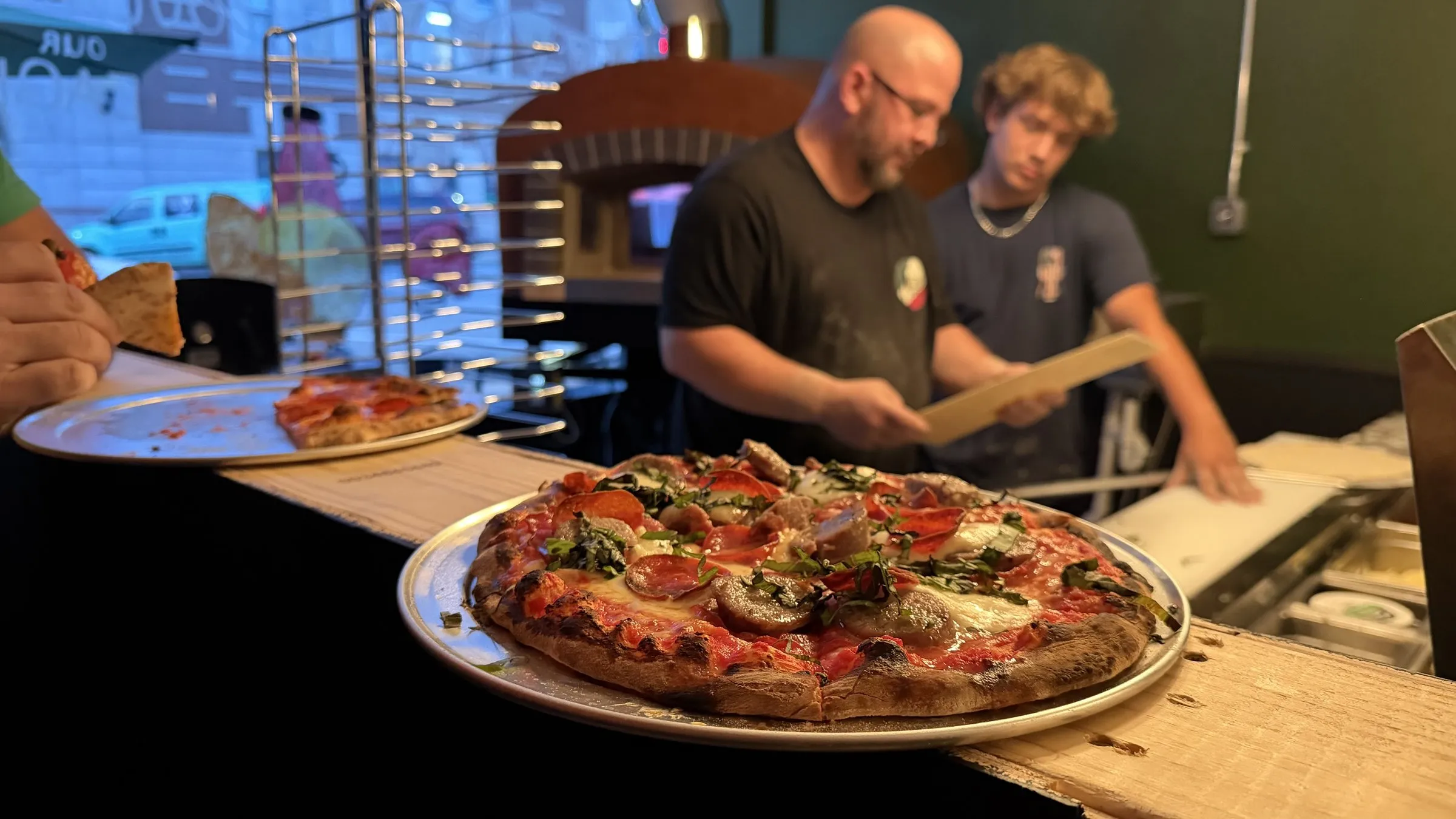 A finished pizza on the counter in front of the wood-fired brick oven, with Wes and a cook working the line behind.